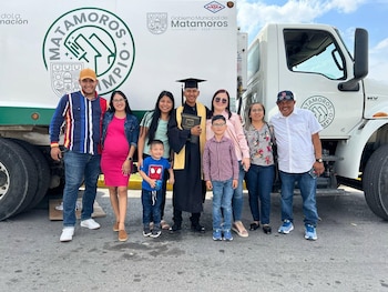 Un hombre con toga, birrete y estola de graduación sostiene su título junto a un camión municipal blanco con la puerta abierta