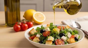 Aceitera vierte aceite sobre ensalada con tomate, aceitunas, rúcula, queso feta y croutones en tazón blanco. Botella de aceite, limones y cubiertos sobre mesa.