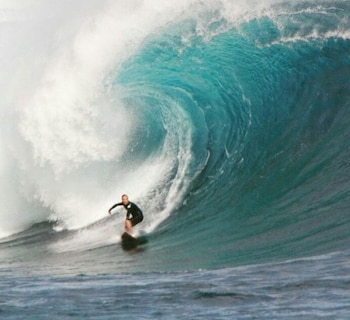 Un hombre con traje de neopreno negro sobre una tabla de surf oscura se desliza por una gran ola turquesa que rompe en el océano