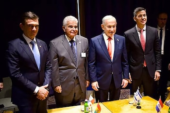 Cuatro hombres con traje oscuro permanecen de pie tras una mesa que exhibe banderas en miniatura de Israel, Paraguay y Panamá