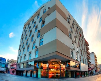 Edificio de varias plantas con fachada beige y blanca, múltiples ventanas, unidades de aire acondicionado, entrada de cristal, banderas, calle y cielo azul