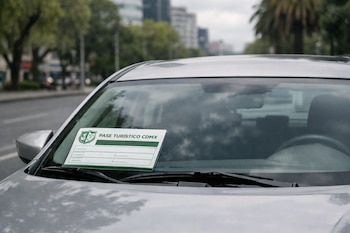 Automóvil plateado con Pase Turístico CDMX visible en el parabrisas. El pase tiene el logo del Gobierno de la Ciudad de México. Fondo urbano desenfocado con edificios y vegetación.