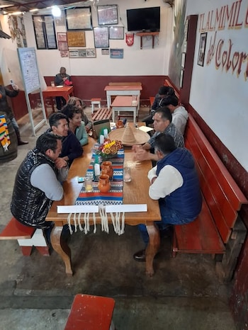 Personas sentadas a una larga mesa de madera con un sombrero de paja, flores y vasijas de barro en un salón con cuadros