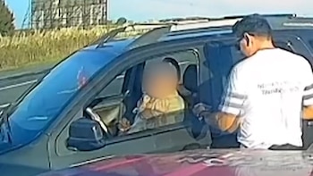 Un oficial de tráfico conversa con un conductor dentro de un SUV oscuro en una carretera. Al fondo se ven plantas y una estructura