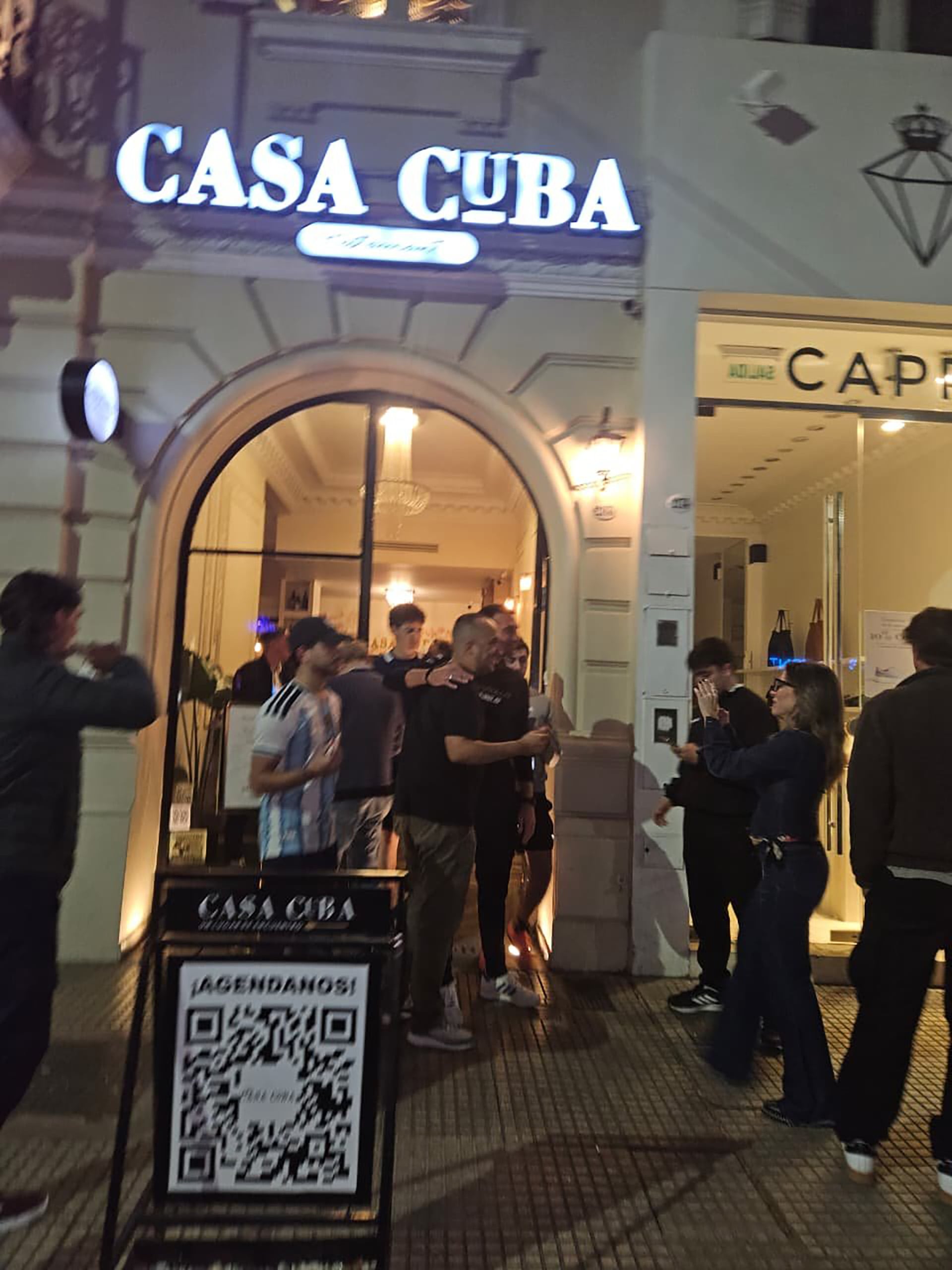 Un grupo de personas conversa y toma fotografías en la entrada del establecimiento Casa Cuba durante la noche.
