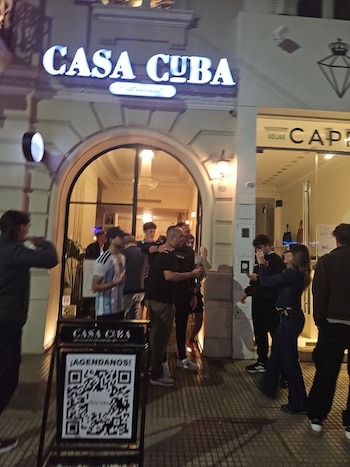 Varias personas en la acera frente al letrero iluminado de Casa Cuba y un cartel con un código QR en primer plano