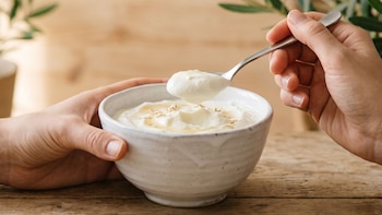 Dos manos sujetan un tazón blanco con yogur griego, miel y avena. Una cuchara de metal levanta yogur. Mesa de madera y plantas verdes difuminadas
