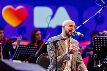 Hombre calvo con barba, anteojos y saco rayado canta en micrófono. Fondo azul con logo de corazón y letras "BA". Orquesta borrosa detrás