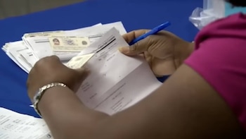 Primer plano de una mano sosteniendo documentos y un bolígrafo azul, con papeles y una tarjeta de identificación sobre una mesa azul