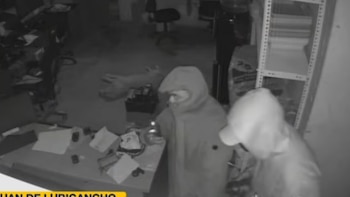 Captura de pantalla en blanco y negro donde dos personas con capucha revisan el área cerca de un escritorio en una oficina