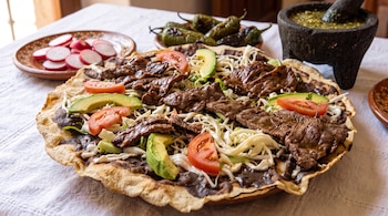 Tlayuda con frijoles, queso, aguacate, jitomate y carne de res sobre un mantel blanco, acompañada de rábanos y salsa verde.