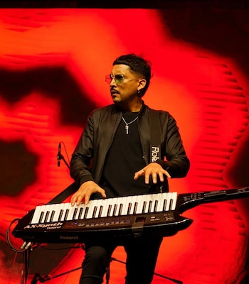 Un hombre con gafas y ropa oscura toca un teclado sintetizador portátil ante un fondo rojo