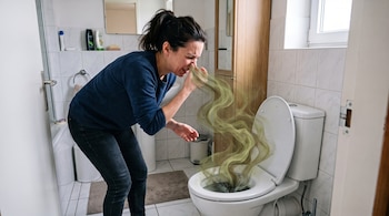Una mujer se tapa la nariz frente a un inodoro abierto del que sale una nube de vapor verdoso.