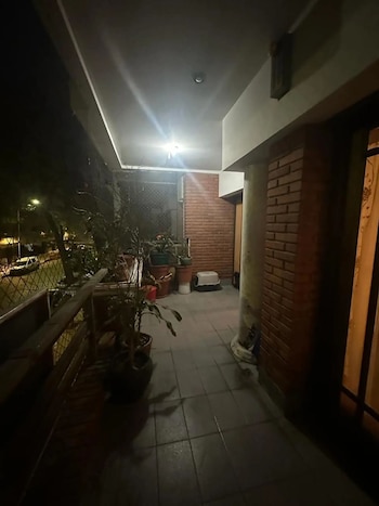 Vista nocturna de un balcón con plantas en macetas, una pared de ladrillos, un pilar, y una red de seguridad. Se ve la calle iluminada a lo lejos