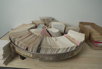 Un contenedor circular gris con tarjetas de índice, rotulado 'NOMINALS A-Z'. Pilas de papeles, una caja de cartón con documentos y una mesa de madera