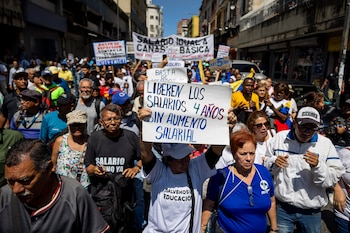 Multitud de trabajadores venezolanos marchando en una calle, portando carteles de protesta con mensajes sobre salarios. Se ven edificios al fondo bajo un cielo azul