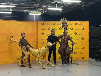 Tres personas de pie ante un fondo amarillo con el logotipo de El Rey León, dos de ellas con vestimentas y marionetas de animales