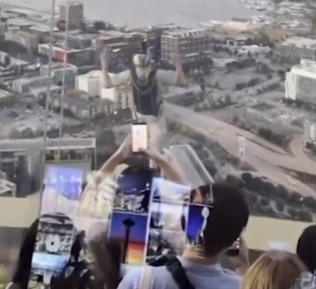 Persona saltando en paracaídas. Vista aérea de ciudad con edificios y cuerpo de agua. Cristales con reflejos de personas y teléfonos móviles
