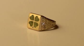 Anillo dorado con un trébol de cuatro hojas verde en el frente, una piedra brillante incrustada y grabados sobre una superficie clara