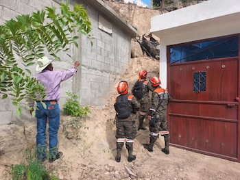 Un hombre y tres rescatistas con cascos observan una casa de bloques de cemento junto a una puerta marrón y un gran deslizamiento de tierra
