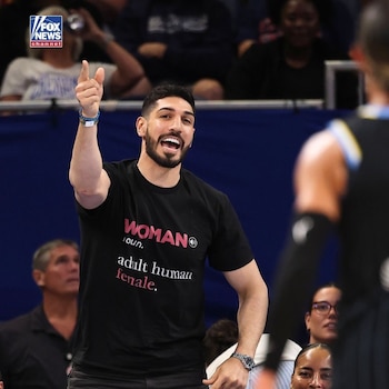Enes Kanter Freedom viste una camiseta negra con texto en inglés y señala hacia arriba con el dedo índice en un pabellón deportivo