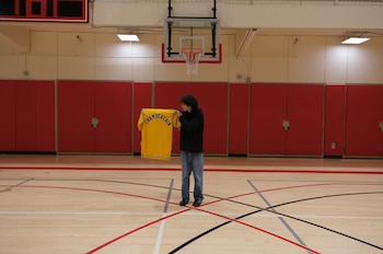 Un adolescente en una cancha de baloncesto sostiene una chaqueta amarilla con el nombre 'CHAMBERLAIN' en azul, debajo de un aro de baloncesto
