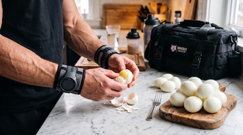 Unas manos con muñequeras pelan un huevo cocido junto a cáscaras, varios huevos pelados sobre una tabla y un bolso deportivo negro.