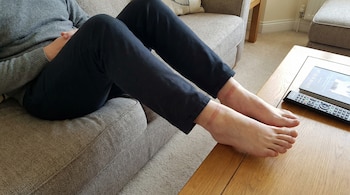 Piernas y pies de una persona sentada en un sofá, con marcas rojizas en los tobillos por medias ajustadas. Se ven pantalones oscuros y una mesa de madera.