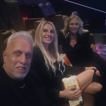 Un hombre de cabello canoso y dos mujeres rubias sentados en butacas de cine con bolsas de palomitas de maíz