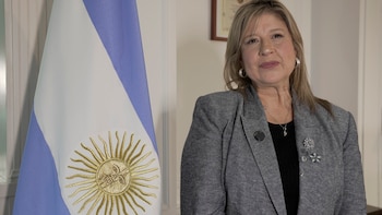 Retrato de una mujer rubia de mediana edad, con blazer gris y blusa negra, de pie junto a la bandera argentina
