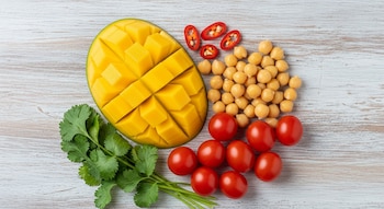 Vista cenital de un mango en cubos, garbanzos, jitomates cherry, cilantro fresco y rodajas de chile rojo sobre una superficie de madera clara.