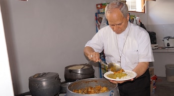 Hombre de cabello gris con camisa blanca y cruz al cuello sirve arroz, guisantes y guiso de carne en plato blanco. Cocina con ollas grandes