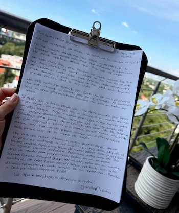 Una mano sostiene una carpeta con una hoja con texto manuscrito y la firma de Sandra Cuevas, en un balcón con plantas al fondo