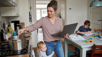 Una mujer con moño cocina en una olla, sosteniendo una laptop con una mano, mientras un bebé rubio se aferra a su pierna y un niño hace tareas en la mesa.