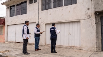 Tres inspectores del SAT con chalecos azul marino y portapapeles esperan frente a la puerta de una casa con fachada blanca y ventanas enrejadas.