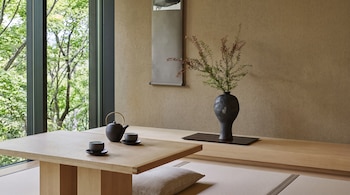 Una habitación con una gran ventana, árboles verdes, una mesa baja de madera, una tetera y dos tazas, un cojín, un jarrón con ramas y un pergamino.