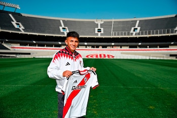 Un hombre joven sonriente de pie en un campo de fútbol verde, sostiene una camiseta de fútbol blanca con una banda roja. Al fondo se ve un estadio con tribunas