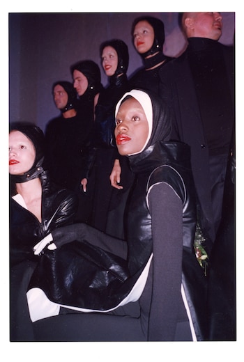 Primer plano de una mujer sentada con capa negra de cuero y capucha con banda blanca, labios rojos, junto a otras personas con vestuario oscuro