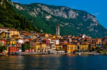 Paisaje de Varenna con edificios coloridos junto a un lago azul oscuro, una torre de iglesia puntiaguda y una gran montaña verde bajo un cielo despejado