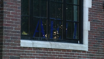 Ventana de marco negro sobre una fachada de ladrillo que contiene letras griegas de color azul pegadas en los cristales