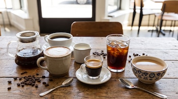 Varias tazas con café, una jarra, un vaso con hielo, granos de café y dos cucharas sobre una mesa de madera.