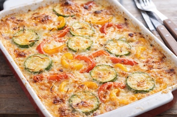 Primer plano de un pastel de calabacín y pimiento gratinado en una fuente de cerámica blanca con borde rojo. Rodajas de calabacín y pimiento cubiertas con queso gratinado.