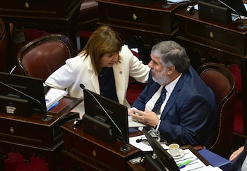Mujer en blazer blanco y hombre en traje azul conversan atentamente en una sala de reuniones con escritorios de madera, monitores y micrófonos
