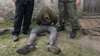 Persona con suéter cubriendo su cabeza yace en el suelo, flanqueada por dos hombres de pie, uno en pantalones negros y otro en camuflaje