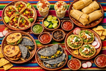 Vista aérea de una mesa con manteles coloridos cargada de platillos mexicanos: tacos, esquites en vasos, tamales, quesadillas azules, gorditas y varias salsas.
