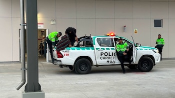 Una camioneta pickup blanca de la policía con luces de emergencia encendidas. Cuatro policías y dos personas civiles cerca de la camioneta. Entrada de un edificio con cartel de 'NCIAS'