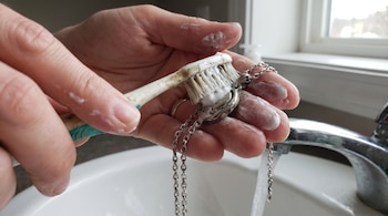 Manos sostienen un cepillo de dientes con pasta blanca limpiando un collar de plata. Hay agua saliendo de un grifo sobre un lavabo blanco.