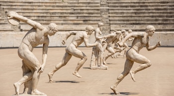 Esculturas de mármol de hombres desnudos en posturas de carrera y lanzamiento de disco sobre una superficie clara con gradas de piedra al fondo