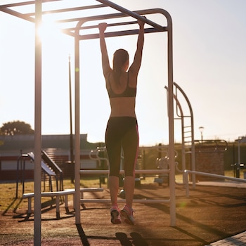 Vista trasera de una mujer en silueta haciendo ejercicio en barras paralelas de metal en un gimnasio al aire libre, con el sol brillante de fondo.