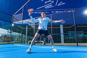 Hombre con chaqueta azul, sostiene raqueta de pádel negra y pelota de tenis amarilla. Mira a la derecha. Detrás, edificio grande y elegante bajo cielo azul